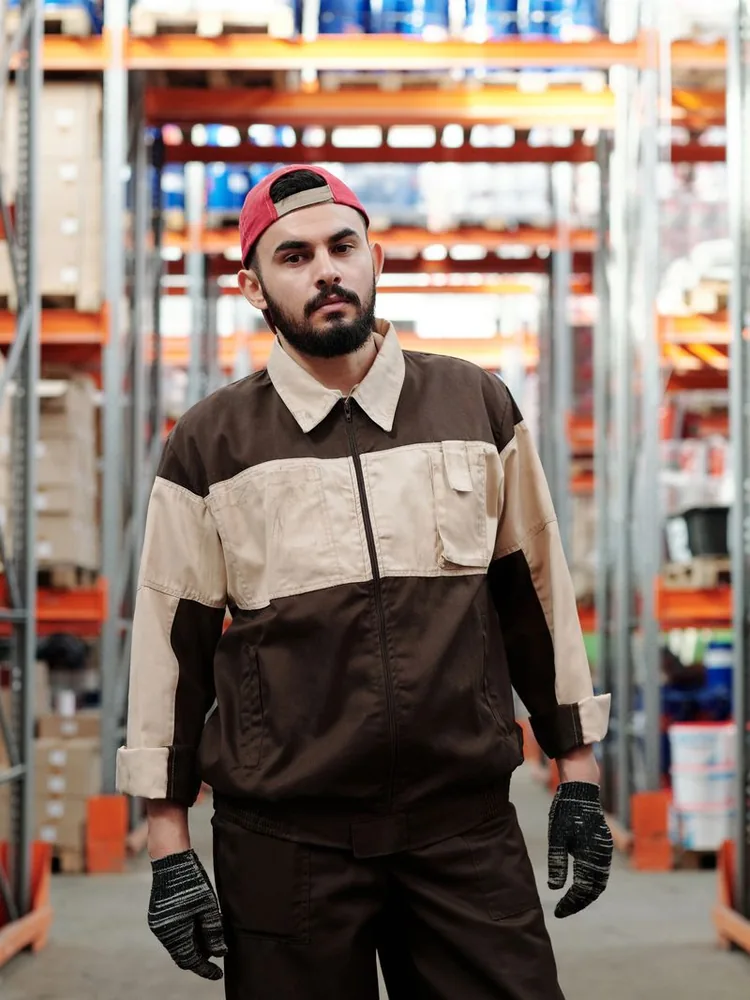Warehouse worker wearing work clothes and gloves in front of storage shelves.